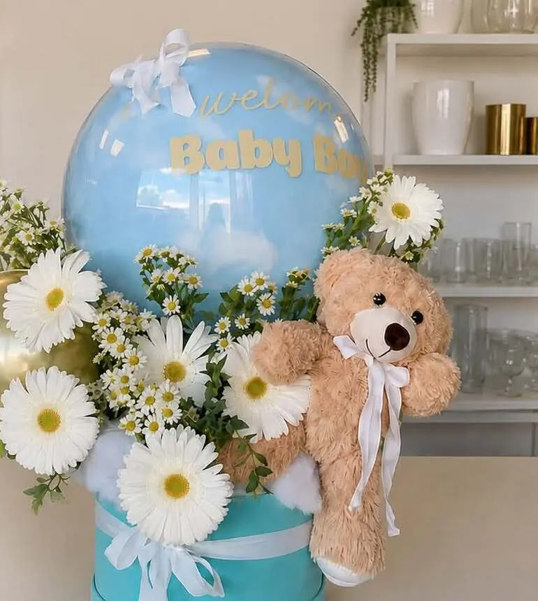 Arreglo de margaritas blancas y un osito de peluche café, con un globo azul que dice “Welcome Baby Boy”.