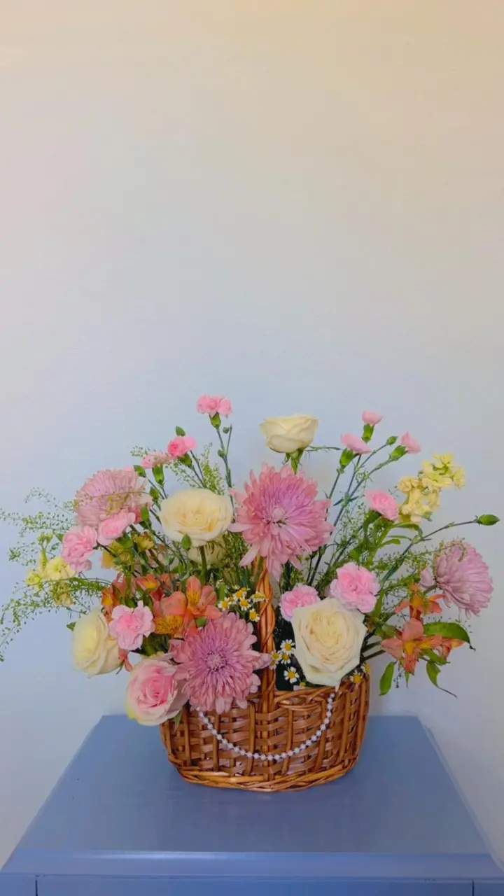 Canasta de mimbre adornada con un hilo de perlas, llena de crisantemos rosados, rosas blancas, claveles y alstroemerias, sobre una mesa azul.