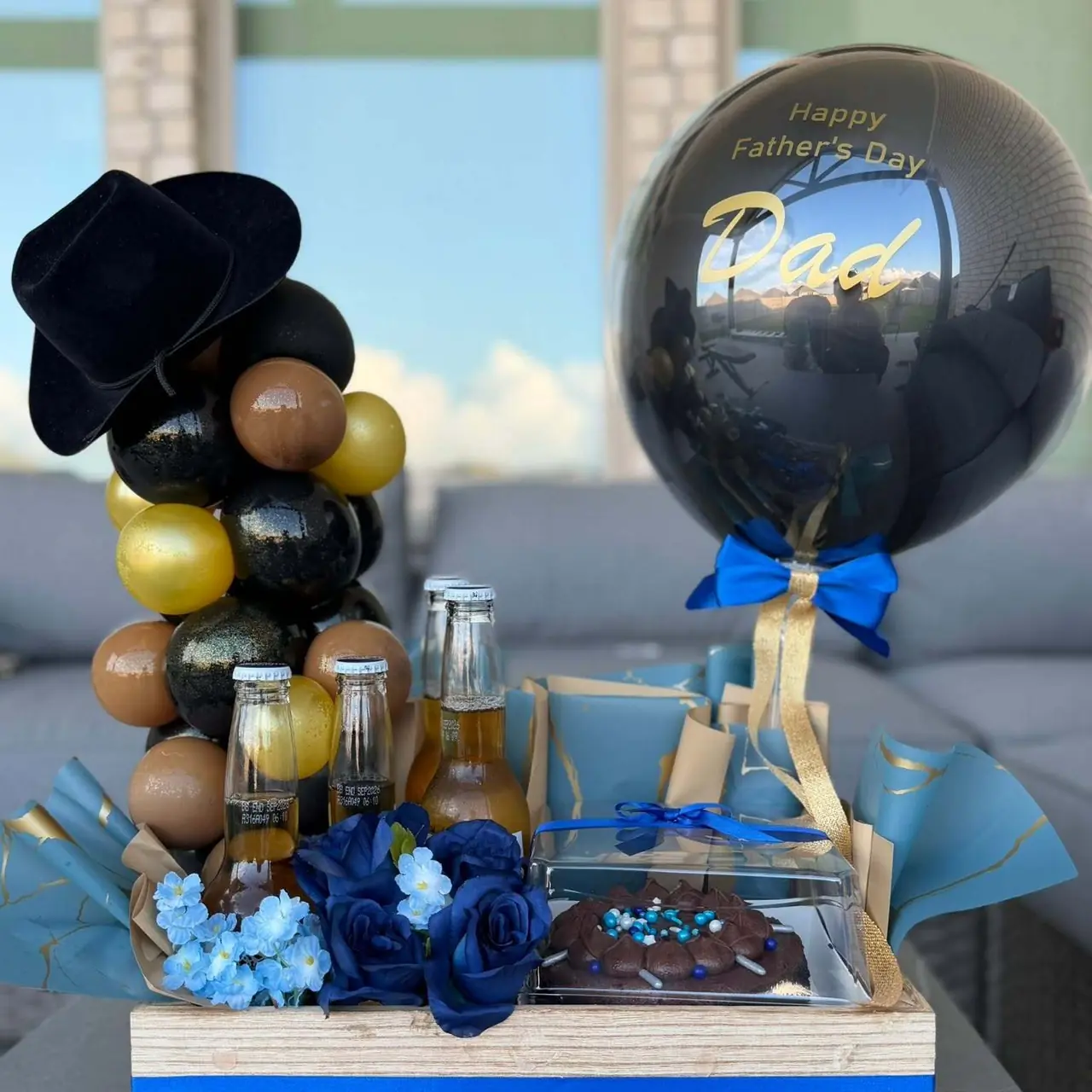 Regalo de Día del Padre: torre de globos negros, dorados y café con sombrero, cervezas, rosas azules, un pastel de chocolate y un globo que dice “Happy Father's Day Dad”.