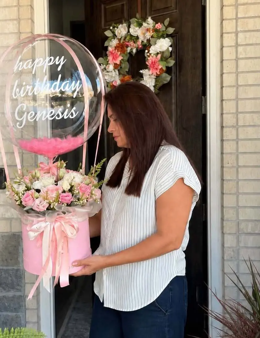 Mayina entregando en la puerta de una casa un arreglo de rosas rosadas en caja con un globo que dice “happy birthday Genesis”.