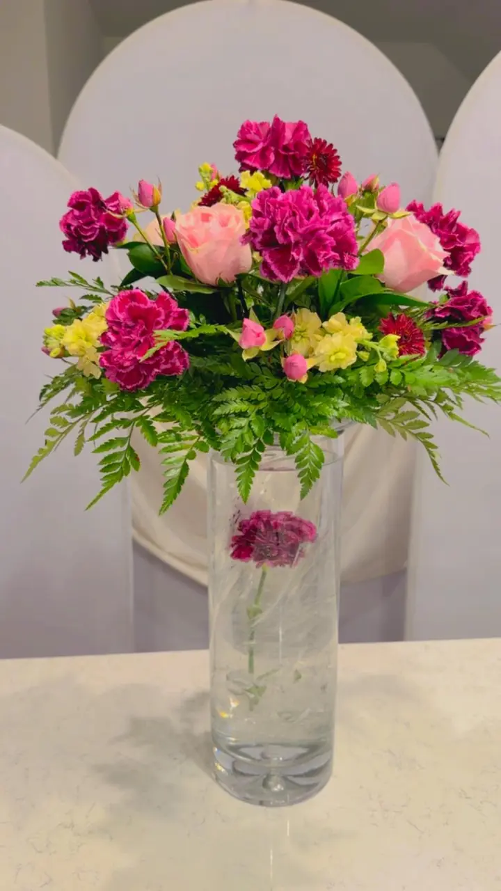 Arreglo de claveles fucsia, rosas rosadas, flores amarillas y helechos, en un jarrón alto de vidrio.