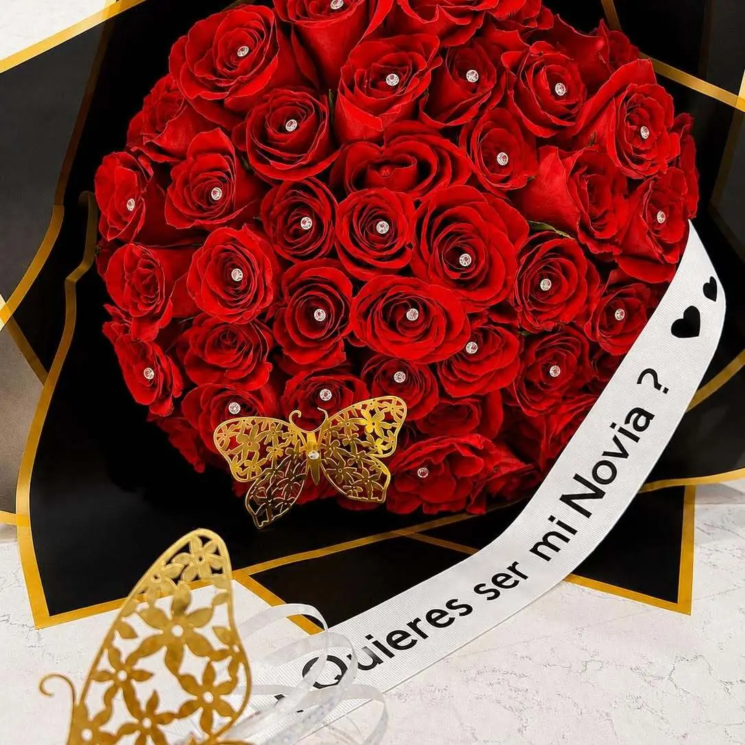 Ramo de rosas rojas con cristales en el centro de cada flor, envuelto en papel negro y dorado, con banda que dice “¿Quieres ser mi novia?” y una mariposa dorada.